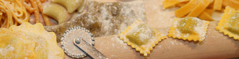 Assorted types of pasta with a rolling pin and pasta cutter on a wooden board.